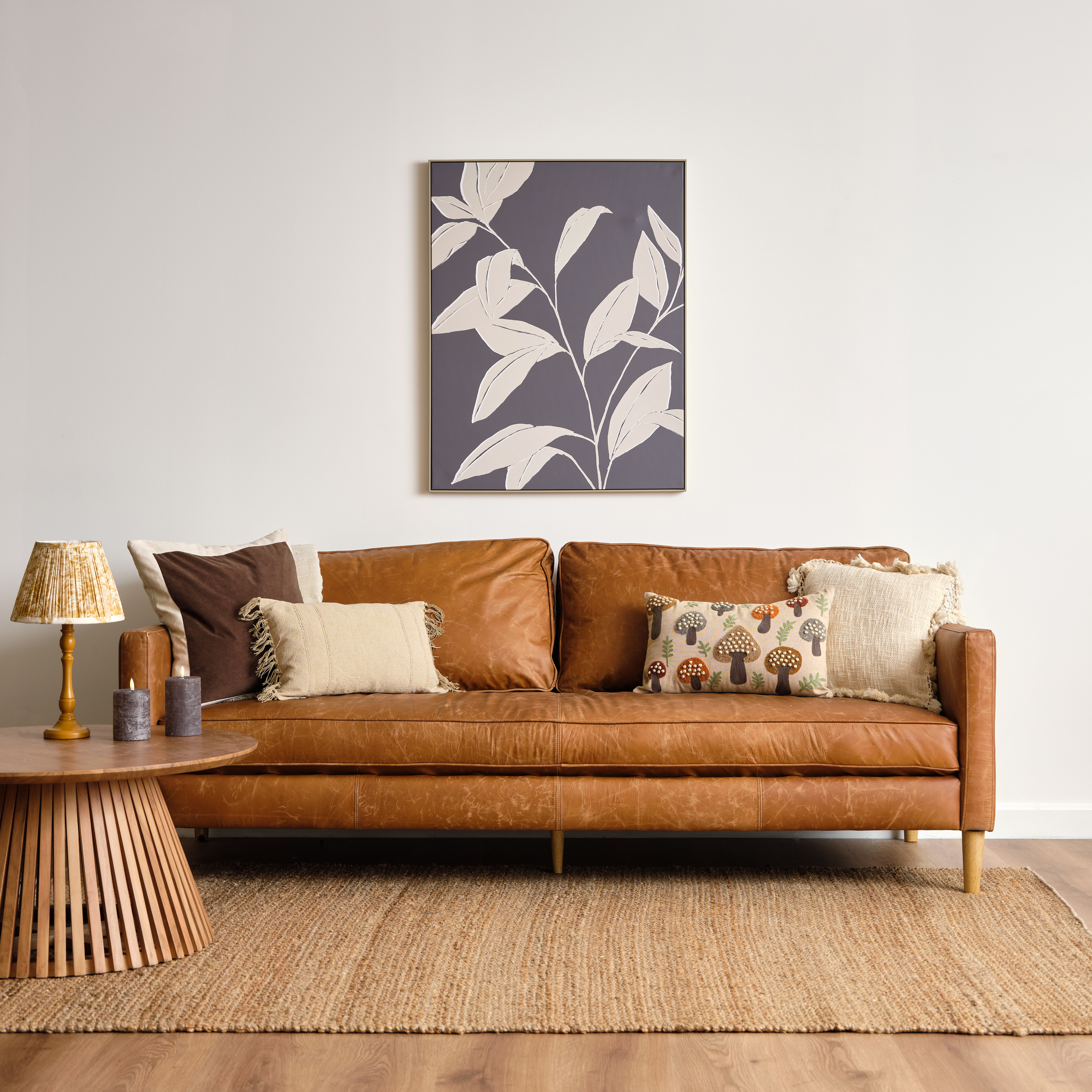 a tan leather sofa in a room with art of foliage hanging above and a side table with a lamp on it