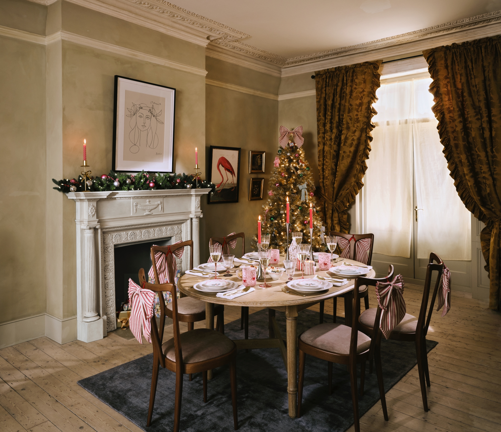 a dining room with red striped bows on the wooden chairs, a christmas tree to the right of the fireplace and a christmas garland on the fireplace mantle