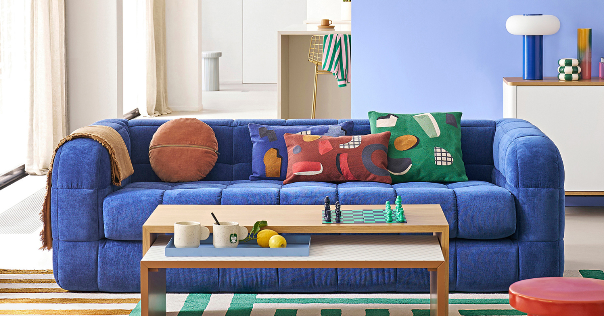 Pieta sofa in blue in a lounge with a light wood coffee table, a red side table, green and white stripe rug, light blue walls and a blue bibbin round mirror and lamp.